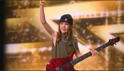 Britain's Got Talent/Freemantle/SYCO Olly with his red electric guitar on Britain's Got Talent. He is holding one hand in the air with a golden background.