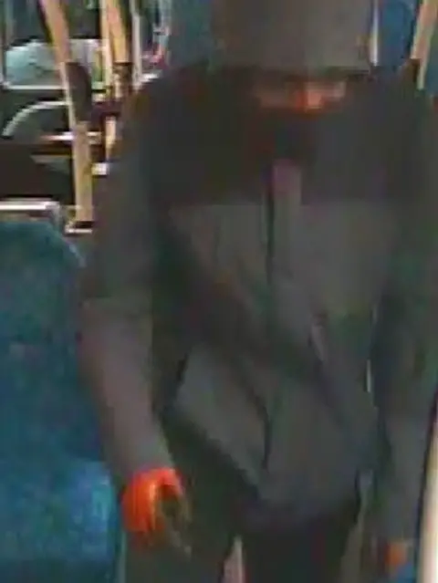 Met Police handout Image of the man wearing a black and grey hooded coat with a red zip and a black facemask, as well as red and black gloves.