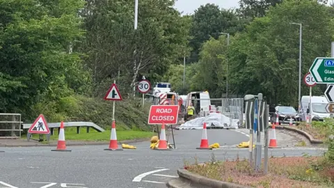 A road closed sign at a roundabout with traffic queued to one side and another area fenced off for work