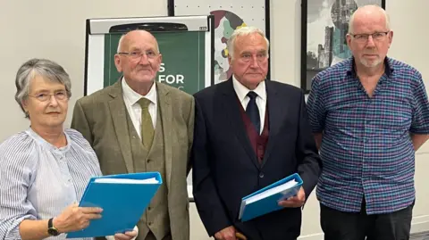 Four people stood in a conference room, two of them are holding blue folders with information inside. All individuals are formally dressed with two wearing shirts and two wearing three piece suits