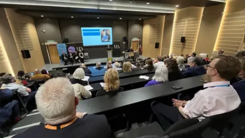 Shaun Whitmore/BBC People sitting in a lecture theatre and several people are sitting and standing on stage