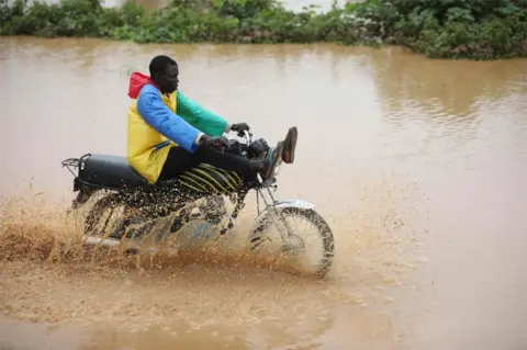 AFP A man wearing a colourful rain-proof jacket is seen riding a motorcycle through brown floodwater. He is balancing his legs close to the handlebars to avoid getting them wet.