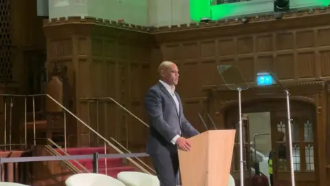 BBC Marvin Rees with a bald head standing at a podium wearing a dark suit and a white shirt addressing a hall