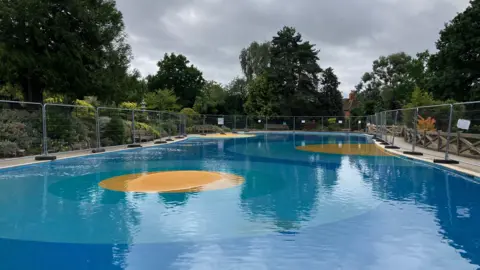 BBC The paddling pool in Stoke Park, Guildford, Surrey, during a period of closure due to water contamination. Wire fencing can be seen around its parameters. 