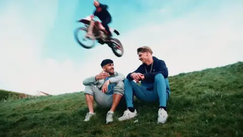 Westroyd Movement A young man on a motorbike flies off the ground, over two young men sat on a grassy hill.