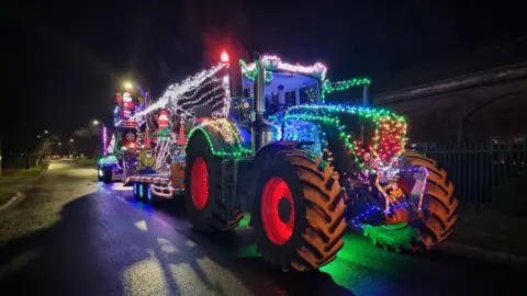 A tractor pulling a trailer, with both covered in lights and decorations. The photograph is taken at nighttime, and there are various different coloured lights covering the vehicle as well as inflatable Santas and Minions characters adorning it.