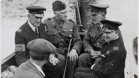 Five men, four wearing uniforms of the British Armed Forces sit in a small boat on the water. The fifth man in the black and white image has his back to the camera and is wearing a jacket with a white shirt collar visible and a flat cap.