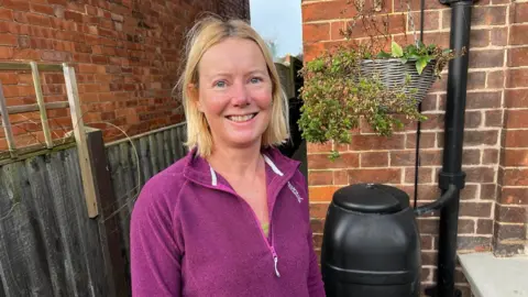 Phil Harrison / BBC A blonde woman in a purple fleece stands in front of a water butt on the side of a house.