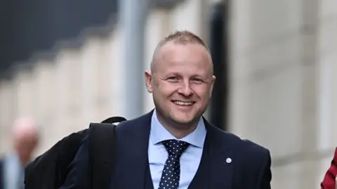 PA Images A man with very short light hair, smiles as he walks towards the court. He wears a navy suit, blue shirt and navy and white polka dot tie. He has a backpack over his right shoulder.
