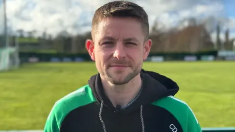 A man with short brown hair and facial hair. He is wearing a green and black hoodie. Behind him is a pitch which is blurred.