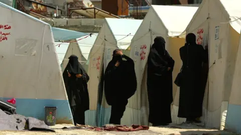 Women wearing face veils stand in front of a row of tents