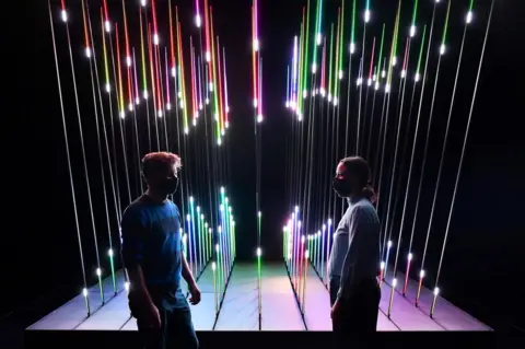 DYLAN MARTINEZ/Reuters Two people wearing face masks stand in front of an illuminated, multicolour exhibit