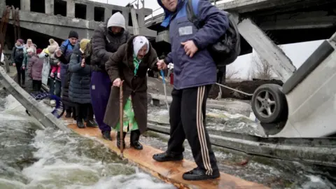 Orla Guerin/BBC A line of people, including children being carried and an elderly woman with a walking stick, gingerly cross the river amid the wreckage of a broken bridge, walking on planks