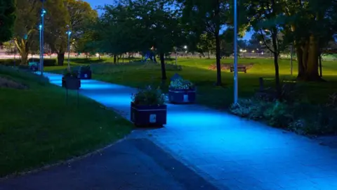 Coventry City Council A footpath in a park lit up in sky blue lighting. Benches, trees and lamposts can be seen in the background.