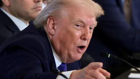 Donald Trump pointing towards the camera, sat next to other people whose faces are not in full view. He is wearing a purple tie and a blue jacket.