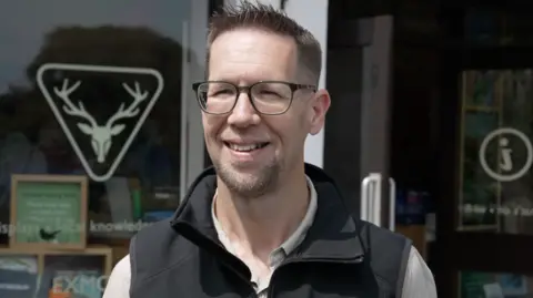 A man wearing a gilet and glasses smiles at the camera outside the National Park Centre in Dulverton