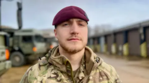 Young man wearing Army fatigues and burgundy beret poses in front of tank-like vehicles