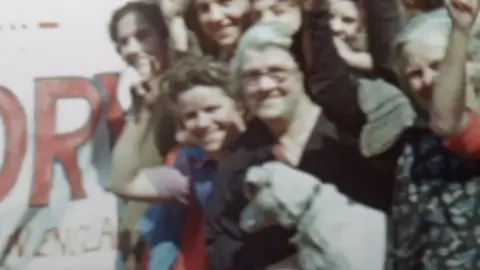 Yorkshire & North East Film Archive Aileen Crooks stands among the crowd and besides a victory banner. She is standing behind her mother, who is holding their family dog.