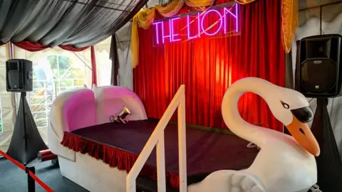 Zac Humpage A swan boat wit its seating area covered in a platform draped in red tasselled material. A red curtain backdrop has a neon pink The Lion sign lit up. Grey material drapes down from the ceiling. The swan is behind a red cordon. Speakers can be seen either side of the stage.