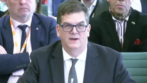 Sir Olly Robbins appearing before the Foreign Affairs Committee. He wears a black jacket, white shirt and black spotted tie.