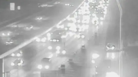 A black and white CCTV still of the overturned car. It has a red square superimposed around it and is lying on one of the road's two lanes. Emergency workers in hi-vis clothing are nearby, as are several of their vehicles.