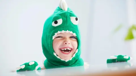A young child wearing a green dinosaur outfit is smiling at the camera.