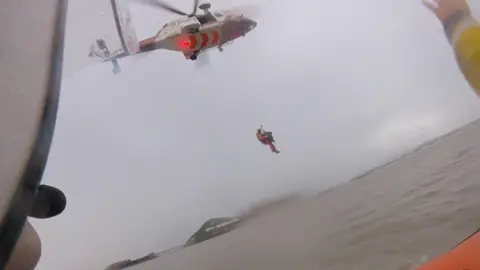 RNLI/Holly Turner A coastguard crew member pulls Mr Grant into a helicopter to transport him safely to hospital. The scene can be seen from the lifeboat. Both the water and the sky are gray.