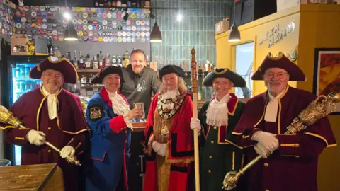 A group of council dignitaries, all dressed in colourful medieval-style robes and three-cornered hats. Two are holding large gold maces and the mayor is wearing a large gold chain. They are in a pub with the bar and landlord in the background.