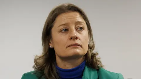 A woman with long brown hair. She is wearing a green blazer over a blue turtle neck jumper. She is also wearing silver earrings.