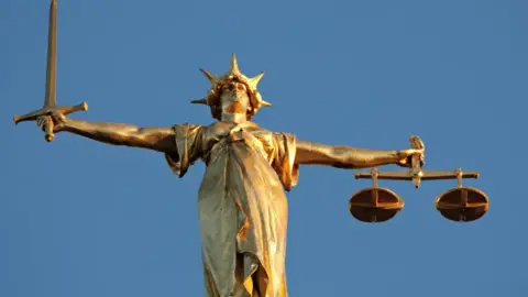 A statue, depicting someone holding a sword in their right hand, in front of a blue-sky background. 