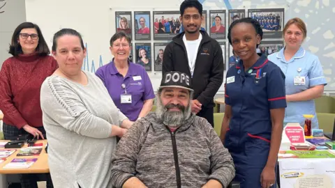 Royal Papworth Paolo Custodio is in the front of the picture. He is wearing a grey, zip-up hoody and black hat with white writing that reads 'dope'. He has a grey and black beard and moustache. Behind him are members of the smoking cessation team. 