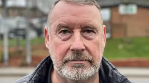 Spencer Stokes/BBC Mick Todd, who has short grey hair and grey stubble. He wears a neutral expression and is staring straight down the camera.