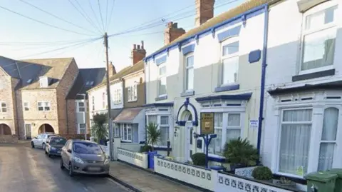 Terraced houses on a residential street, most of them whitewashed. In the centre is a double-fronted former guest house, with two bay windows, an arch above the door and three windows on the upper-floor. The woodwork and other features are painted blue. Cars are parked outside. The road sweeps around to the left, with a more modern red-brick, three-storey building on the corner.