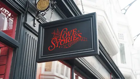 A photo of the Love Stories sign painted in dark grey-green, with red painted lettering. The sign is rectangular and hangs from a wrought iron bracket. The top of the windows can be seen, while above the upper floor windows and the covered skylight of the arcade with metal strut supports can be seen.