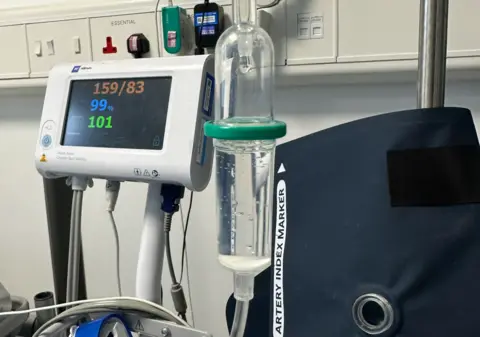 Medical equipment by a wall in a hospital. Electrical sockets are at the top of the image. A screen on the left has numbers in different colours. A plastic or glass bottle-shaped container including liquid is in the middle of the photo.