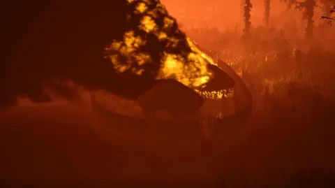 Guy Schofield A digital art installation shows several wooden huts with straw roofs. There is a fence which runs around the huts. A large fire has started in one of the huts and there is a big black cloud of smoke.