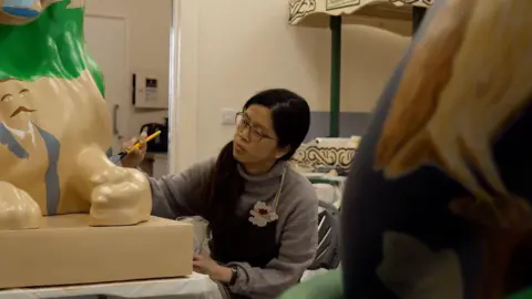 The image shows November Tan sat painting her lion. She has a grey, high neck, knitted jumper on. She has black, long hair which is in a ponytail. She has a black apron on and brown glasses. She is holding a yellow paintbrush. She is painting a brown lion, which has green and grey on it.