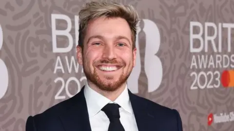 Sam Thompson smiling at the camera. He has blonde hair. He is wearing a white shirt, black tie and jacket. 