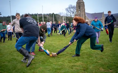 Brook Wassall A woman with a homemade stick competes with two other players for the ball. The Royal Chapel of St John's can be seen in the background.