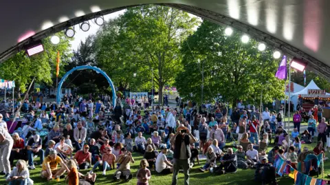 Still Moving Media A large crowd of people sitting in gardens at the jazz festival. The image is taken from the inside of a stage and there are other tents and bunting behind