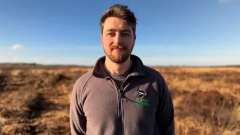 Mr Devenney is looking at the camera. He has a plain expression on his face. He has brown hair and a brown beard and is wearing a branded Ulster Wildlife black fleece.