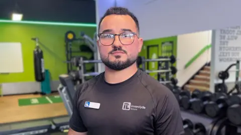 Mo is wearing an Inverclyde Leisure shirt standing in a gym with weights and exercise machines behind him.