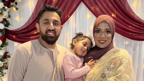 Akeel Nanabawa and Hannaa Vorajee hold their daughter Sara at a celebration event. Akeel wears a grey top while Hanaa wears a terracota head scarf and gold top. Sara is dressed in pink and they stand in front of a floral display