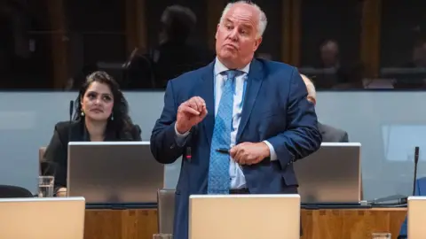 Senedd Cymru Andrew RT Davies, wearing a blue suit, tie and shirt, asking a question in the Senedd chamber watched by Conservative colleague Natasha Asghar