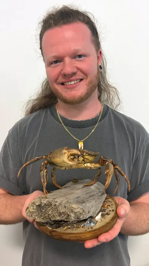 Jason Philip - man with long hair, smiling, holding a crab taxidermy on a plinth