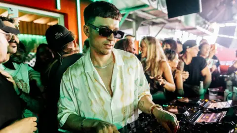 Michael Bibi DJing with people dancing in the background. He is wearing a white shirt and dark sunglasses and is looking down at the DJ decks