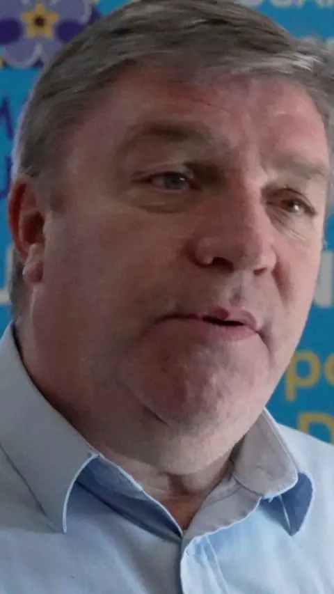  A man with grey hair and eyebrows, talking to the camera. He wears a blue shirt.