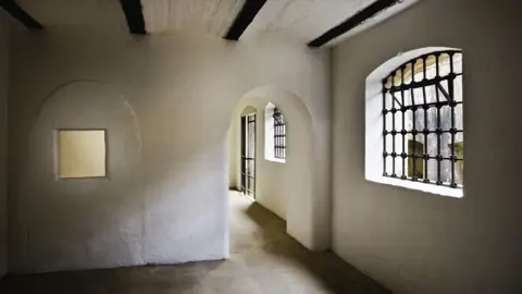 National Trust An empty white plastered room with grey flagstone floor. To the centre left is a square opening in the wall. Centre right is an archway. On the right hand side is a leaded arched window. Black wooden beams cross the ceiling.