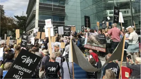Many protestors outside the county council's headquarters, holding placards. Some say "Stop! Highway to hell."