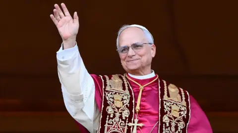Pope Leo XIV wearing a purple shoulder cape that is attached to a white cassock. He wears thin, framed, rectangular glasses and has grey hair. He is waving and smiling slighting looking on from a balcony 